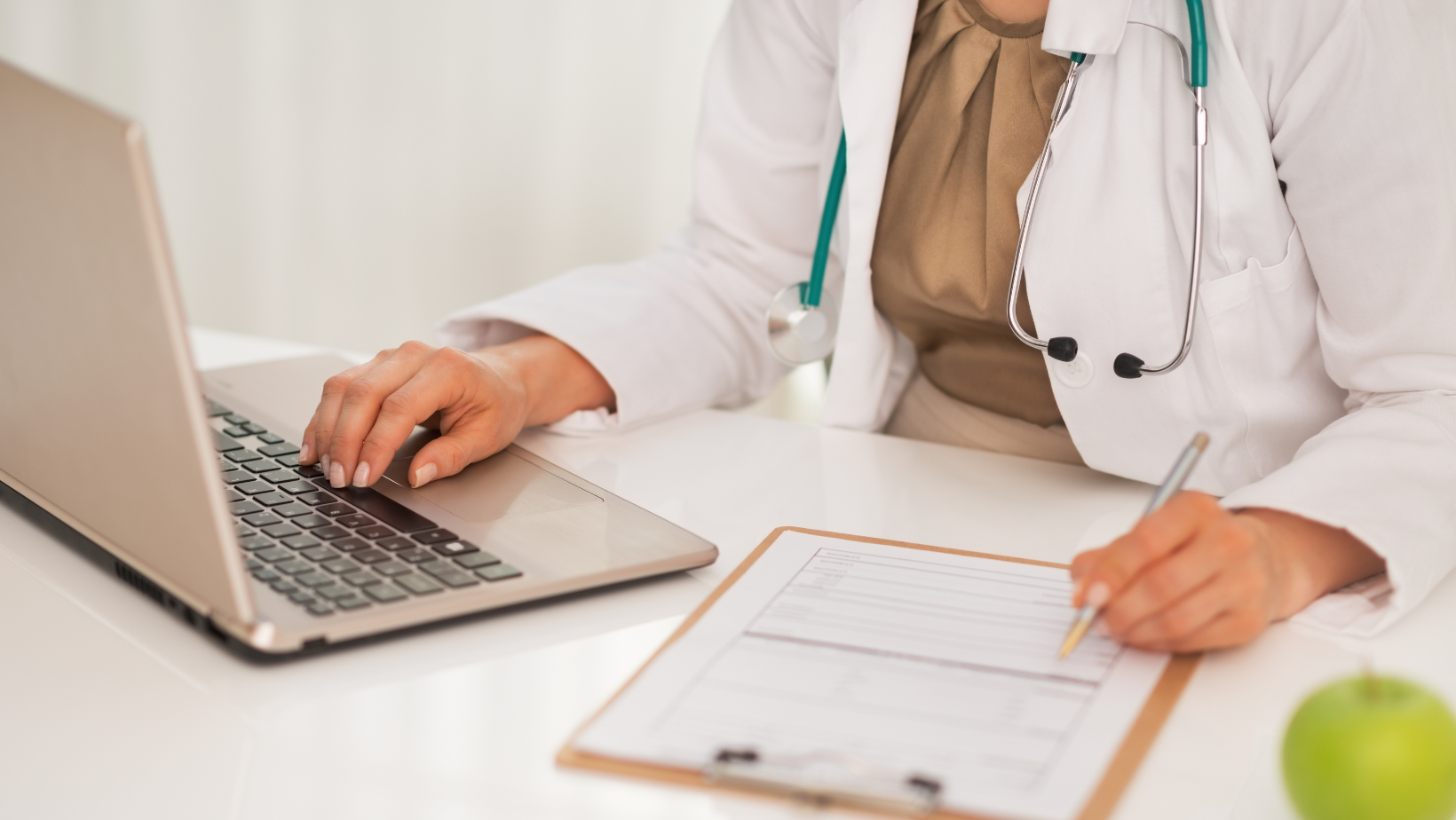 Woman in a white coat with a stethoscope working on a laptop computer with a clipboard